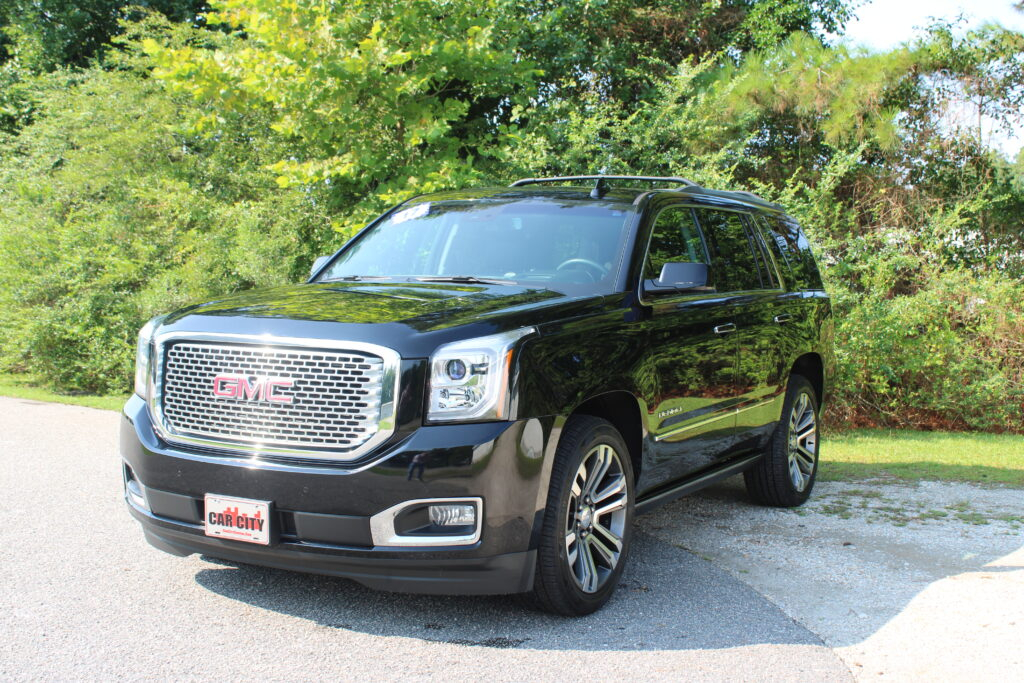 Black GMC SUV parked on a driveway with a chrome grille, visible front bumper, and green bushes behind it. Car City license plate is in view.