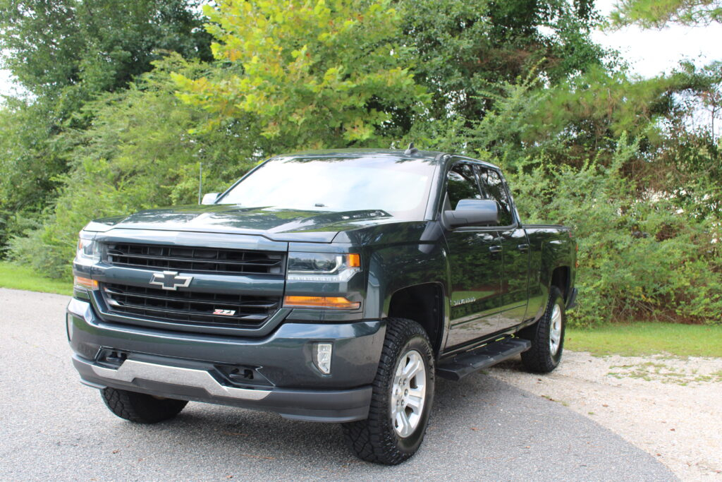 IMG_4353 - Car City Services Front three-quarter view of a dark gray Chevrolet Silverado pickup parked on a driveway with green trees behind it.