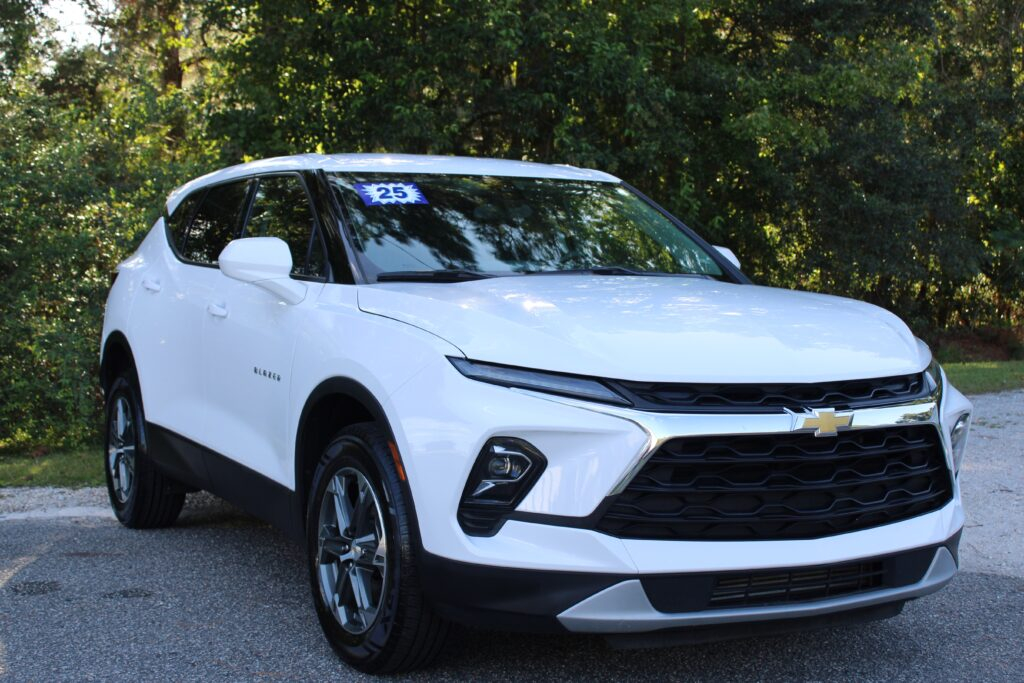 2025 Chevrolet Blazer Stock #39606 - Car City Services White Chevrolet Blazer SUV with a blue '25' sticker on the windshield, parked on a shaded roadway with trees in the background.