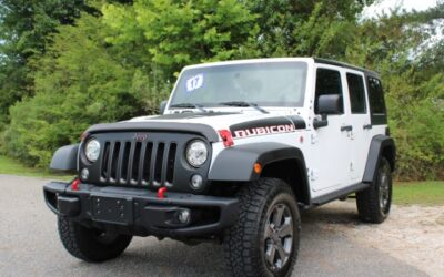Front three-quarter view of a white Jeep Wrangler Rubicon with lifted tires and red recovery hooks, parked on a gravel surface with green trees in the background