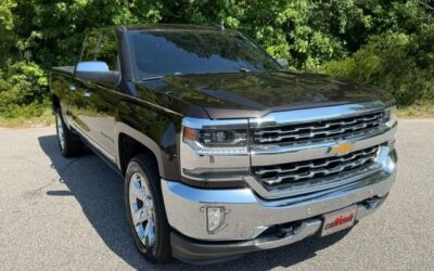 Black Chevrolet Silverado pickup truck parked on a sunlit road with trees in the background, chrome grille and wheels visible.