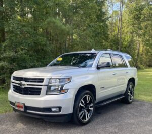 White Chevrolet Suburban SUV parked on a driveway with trees in the background; features black grille and large alloy wheels.