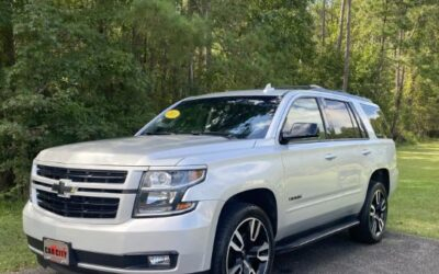 White Chevrolet Suburban SUV parked on a driveway with trees in the background; features black grille and large alloy wheels.