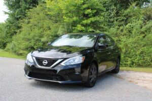 Front three-quarter view of a black Nissan sedan parked on a paved roadside with green trees in the background.