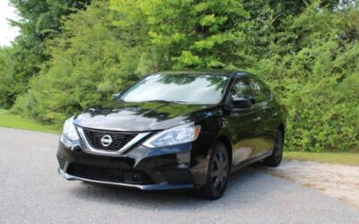Front three-quarter view of a black Nissan sedan parked on a paved roadside with green trees in the background.