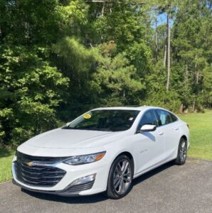 White Chevrolet sedan parked on a paved surface with green trees in the background, sunny day.