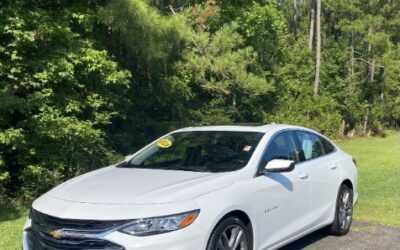White Chevrolet sedan parked on a paved surface with green trees in the background, sunny day.